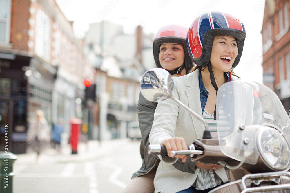 Smiling young women friends wearing helmets, riding motor scooter on urban street Smiling young women friends wearing helmets, riding motor scooter on urban street