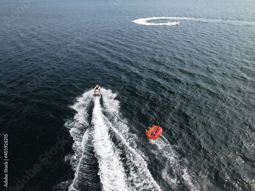 Happy people are going to swim on an air mattress behind a jet ski. Tourists ride the inflatable watercraft boat. Sea attraction. Jet ski skating people on a large Inflatable raft. Aerial copter view.