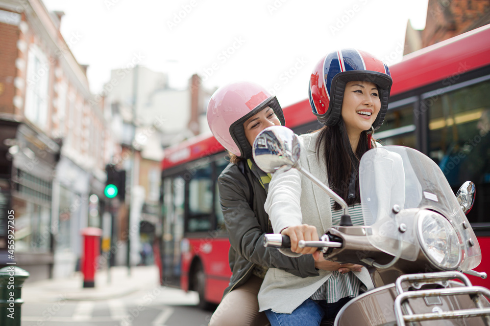 Smiling young women friends wearing helmets, riding motor scooter on urban street Smiling young women friends wearing helmets, riding motor scooter on urban street