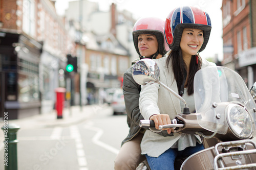 Smiling young women friends wearing helmets, riding motor scooter on urban street