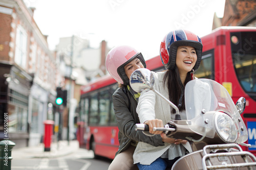 Smiling young women friends wearing helmets, riding motor scooter on urban street