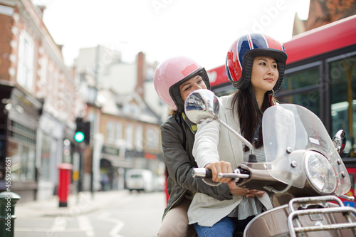 Smiling young women friends wearing helmets, riding motor scooter on urban street