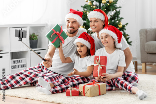 family  winter holidays and people concept - happy mother  father and two daughters in santa hats sitting under christmas tree at home and taking selfie with smartphone