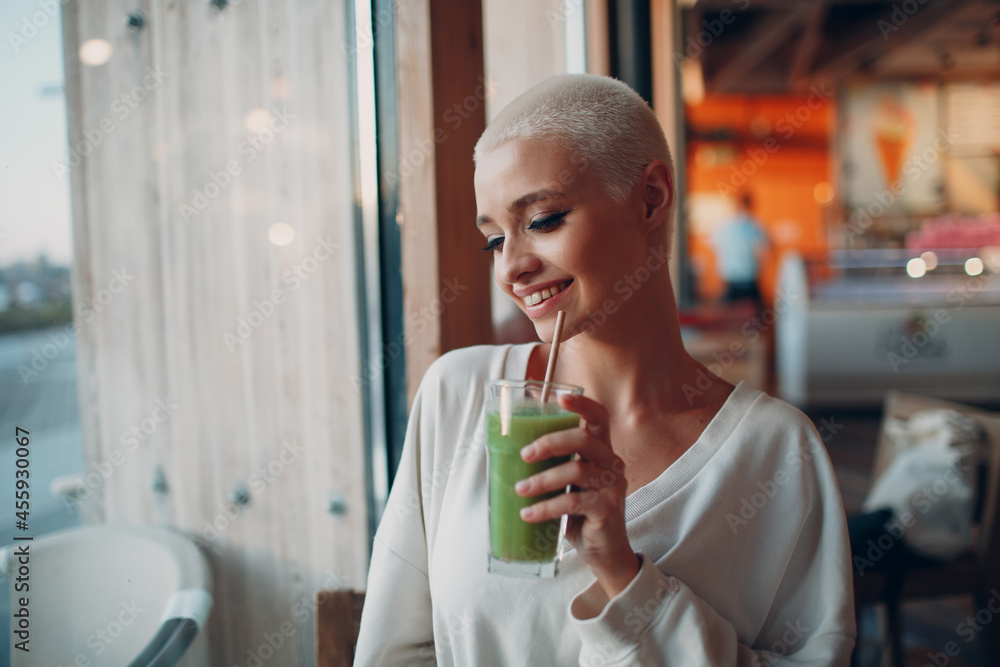 Portrait of young smiling millenial european short haired woman with green smoothie at cafe. Beautiful happy blonde girl indoor. Portrait of young smiling millenial european short haired woman with green smoothie at cafe. Beautiful happy blonde girl indoor.