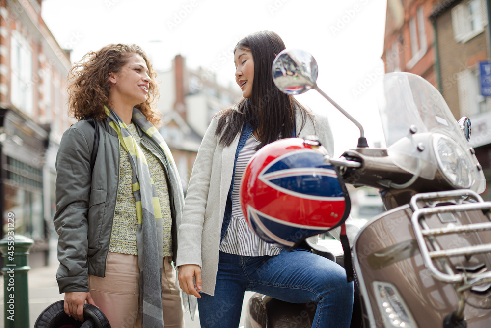 Smiling young woman in helmet on motor scooter, talking to friend on urban street