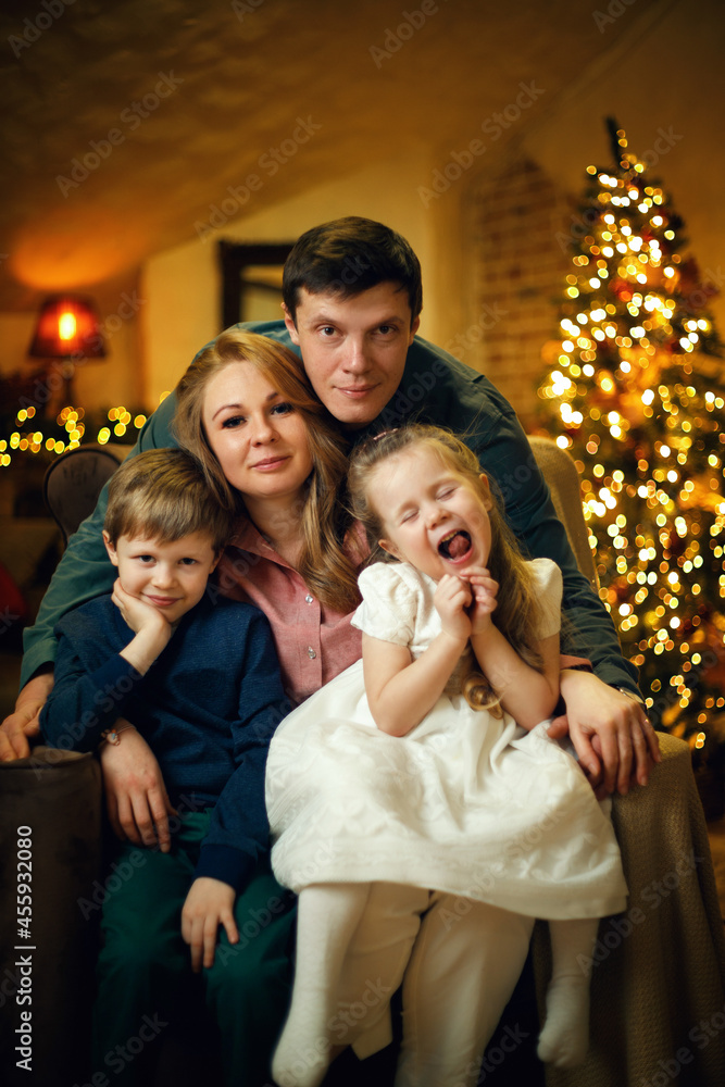 Young beautiful couple with two children posing in a chair in a Christmas interior with a christmas tree and garlands. Selective soft focus, film grain effect