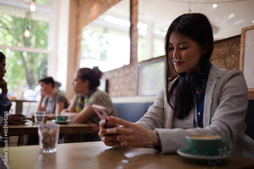 Young woman with headphones texting with cell phone and drinking coffee at cafe table