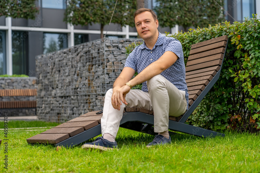 Obraz premium Portrait of smiling handsome millennial man. Positive mature man looking at camera, outdoor backyard at office building recreational area. Handsome man relaxing on wooden chaise lounge in city park.