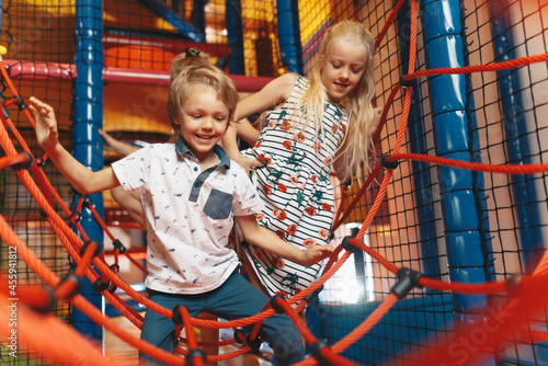 Happy group of siblings playing together on indoor playground. Excited kids playing together on net ropes. Cute school kids playing on colorful playground at shopping mall photo