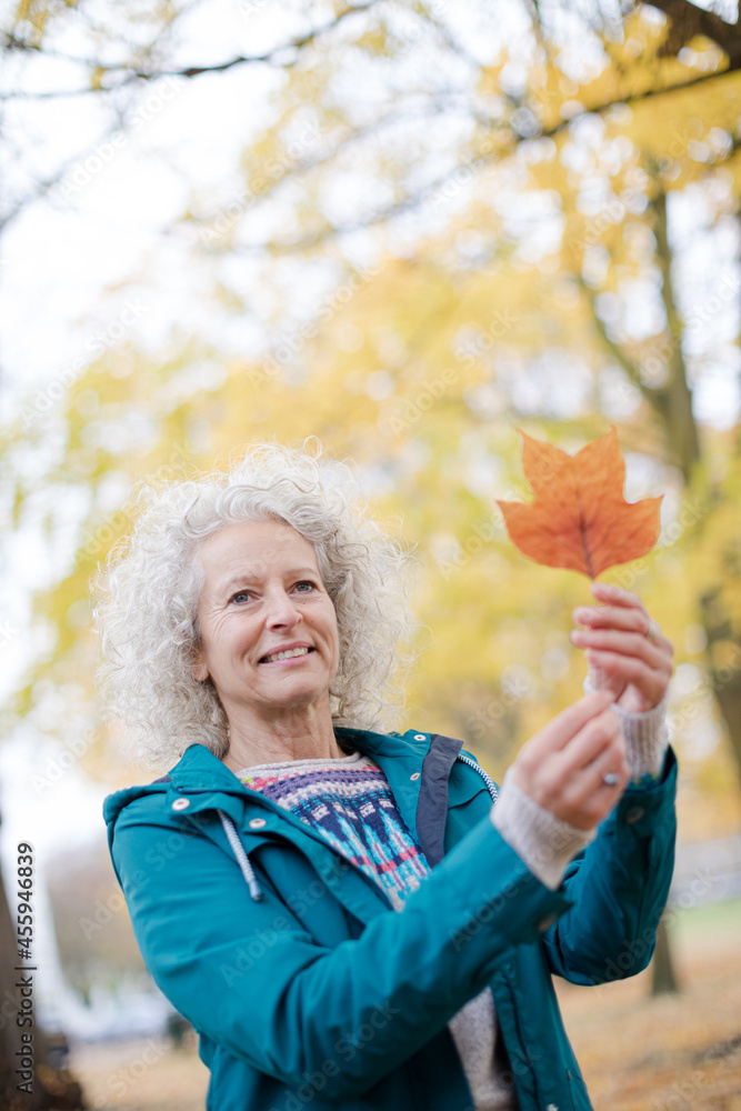 Curious senior woman holding orange autumn leaf in park