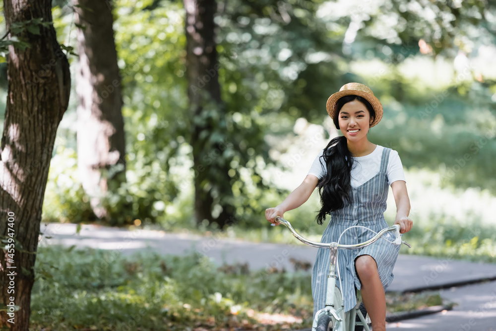cheerful asian woman in straw hat smiling at camera while cycling in park cheerful asian woman in straw hat smiling at camera while cycling in park