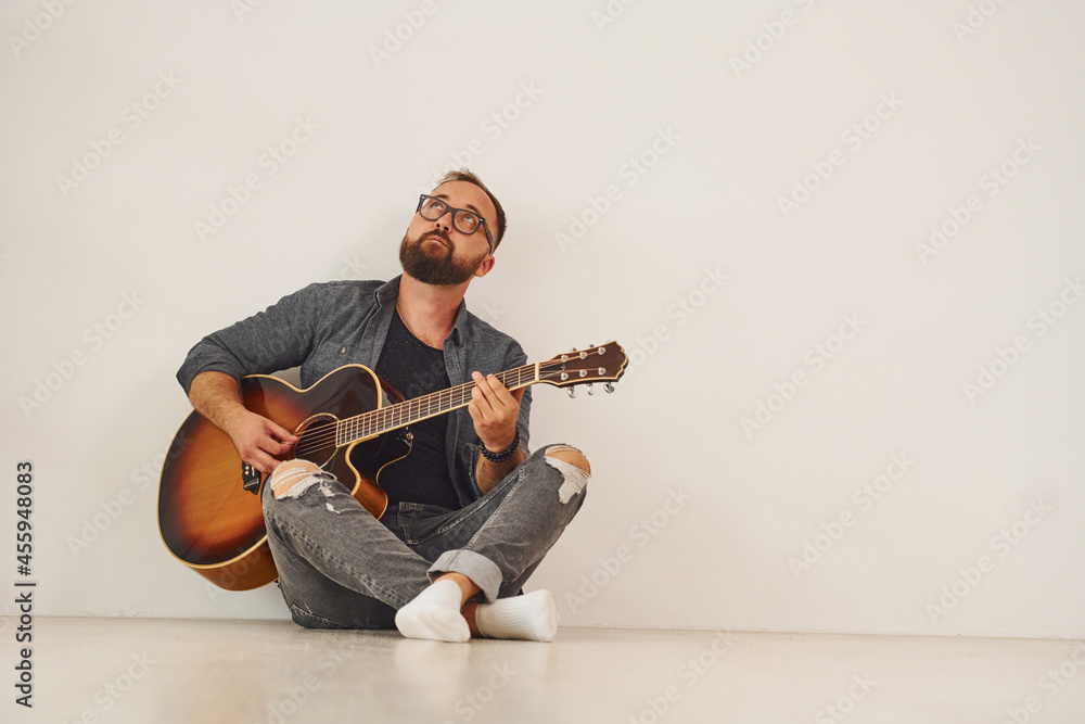 Obraz premium Sitting against wall. Man in casual clothes and with acoustic guitar is indoors