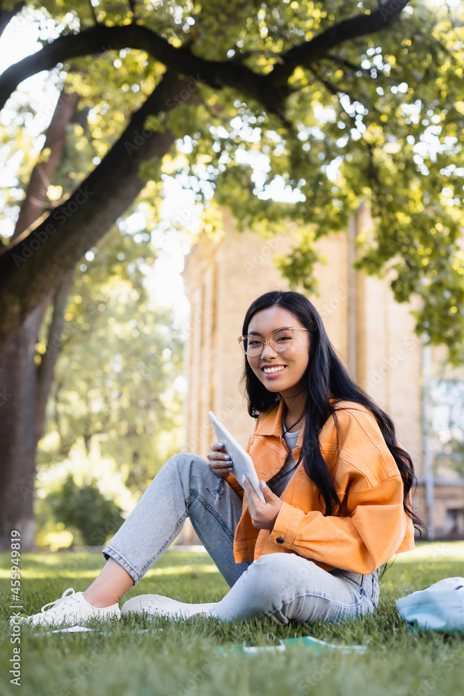 happy asian student in eyeglasses looking at camera while sitting on lawn with digital tablet happy asian student in eyeglasses looking at camera while sitting on lawn with digital tablet