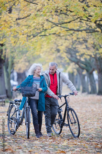 Senior couple walking bicycles among trees and leaves in autumn park