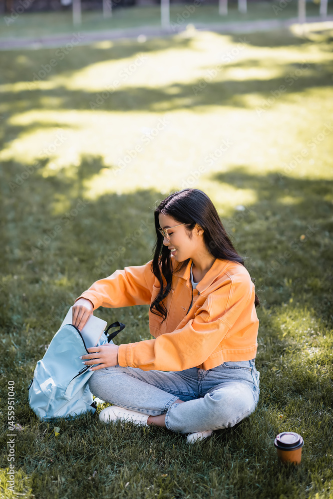 happy asian woman sitting on grass and putting laptop in backpack happy asian woman sitting on grass and putting laptop in backpack