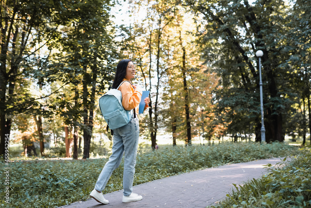 smiling asian woman in jeans walking in park with backpack and notebooks smiling asian woman in jeans walking in park with backpack and notebooks