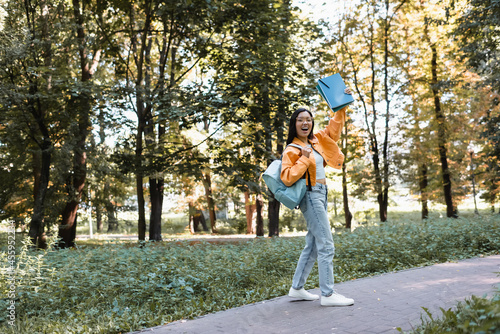 excited asian student waving notebooks while walking in park with backpack photo