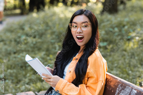 amazed asian student with notebook looking away while sitting on bench in park