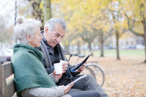 Senior couple reading newspaper and drinking coffee on bench in autumn park