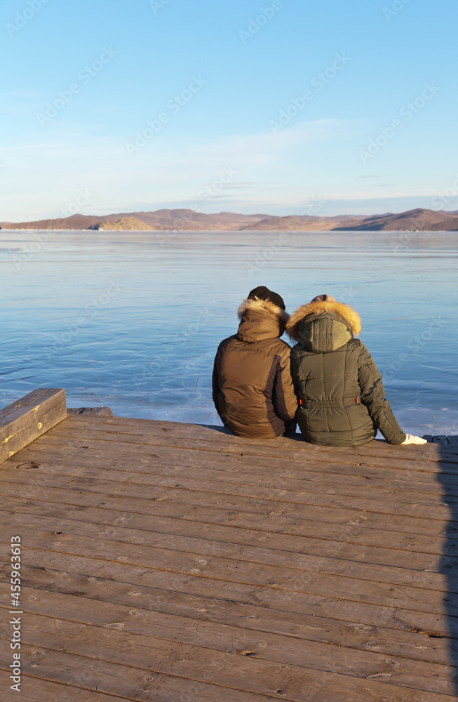 Frozen Lake Baikal in December at sunset. A young couple is relaxing on the wooden pier and admiring the beautiful blue ice in the Kurkut Bay. Winter holidays and travel