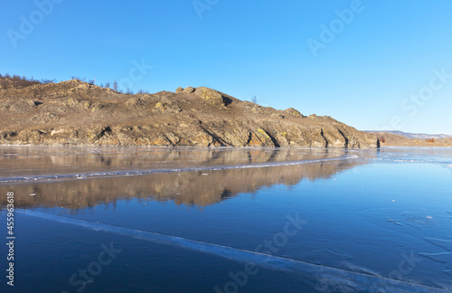 A picturesque winter landscape with a mirror image of coastal rocks in the blue smooth surface of the ice of frozen Lake Baikal on a sunny frosty day. Natural winter background