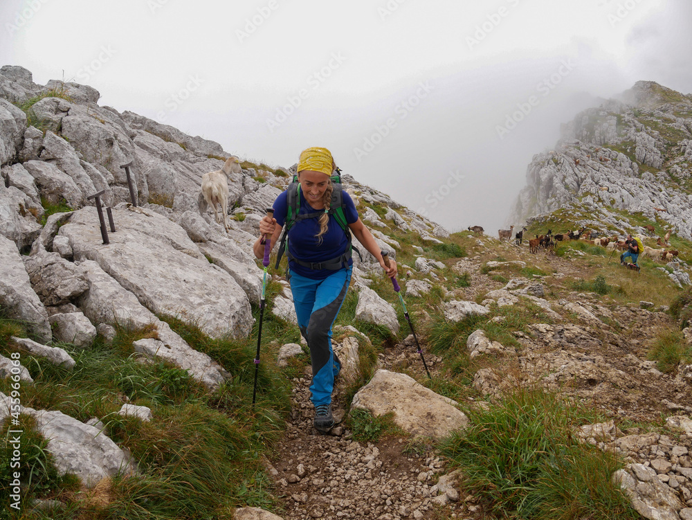 Photo of a Caucasian blonde hiker woman with green backpack, dressed in blue with yellow scarf hiking in a mountainous area with fog