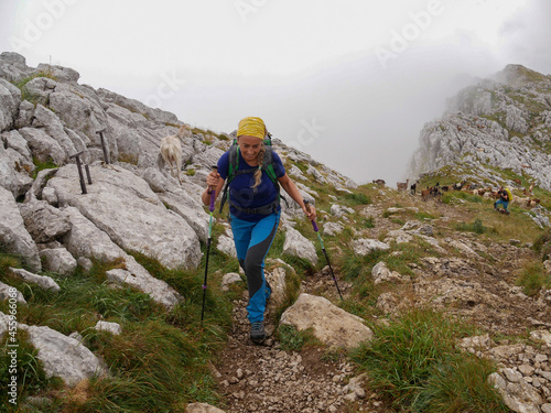 Photo of a Caucasian blonde hiker woman with green backpack, dressed in blue with yellow scarf hiking in a mountainous area with fog