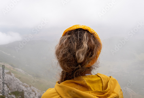 Photo from the back of a Caucasian blonde female hiker, dressed in yellow looking at the landscape in a mountainous area with fog