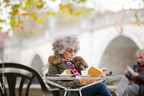 Active senior woman reading book, enjoying cake and coffee at autumn park cafe