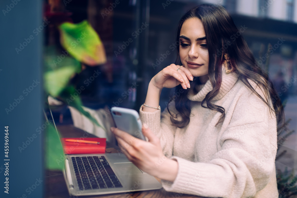 Glad lady using smartphone while working on laptop in cafe Glad lady using smartphone while working on laptop in cafe