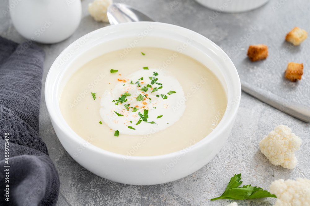 Homemade cauliflower soup puree in a white bowl with cheese, cream, spices and fresh parsley on a gray concrete background. Top view, copy space. Homemade cauliflower soup puree in a white bowl with cheese, cream, spices and fresh parsley on a gray concrete background. Top view, copy space.