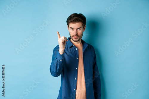 Serious young man scolding person, shaking finger disapprovingly, warn or prohibit something bad, standing on blue background photo