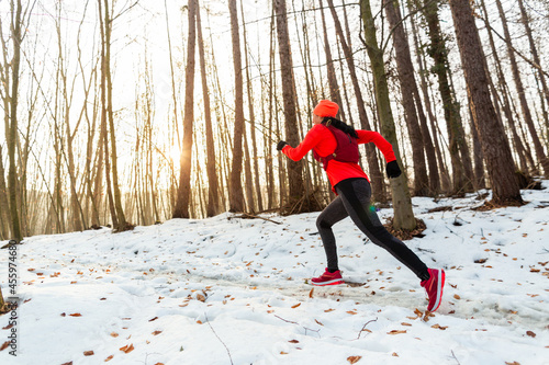 Woman Running Through Woods in Winter. Side View of Female Trail Runner Wearing Hydration Vest Running Uphill in Forest on Cold Winter Day.