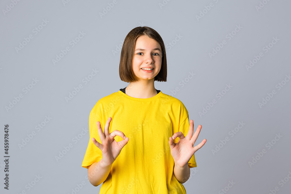Smiling teen girl in yellow casual tshirt show okay sign, praise something impressive, approve and like, stand over light grey background. Happy cute young girl showing okay Smiling teen girl in yellow casual tshirt show okay sign, praise something impressive, approve and like, stand over light grey background. Happy cute young girl showing okay