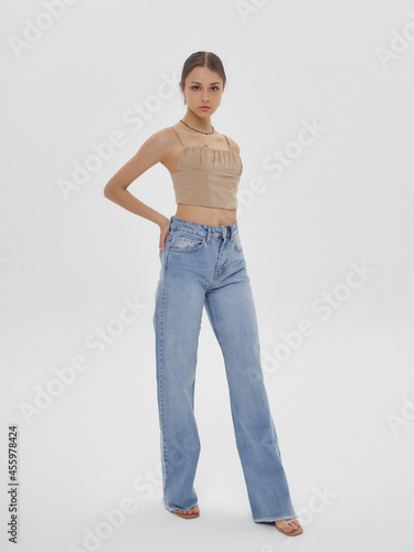 Young elegant woman with long straight hair and natural make-up in beige top and blue jeans standing and posing on bright grey background
