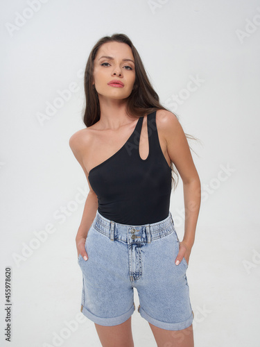 Young elegant stylish woman with long brunette hair and natural make-up in black top, blue shorts and slippers posing against bright grey background