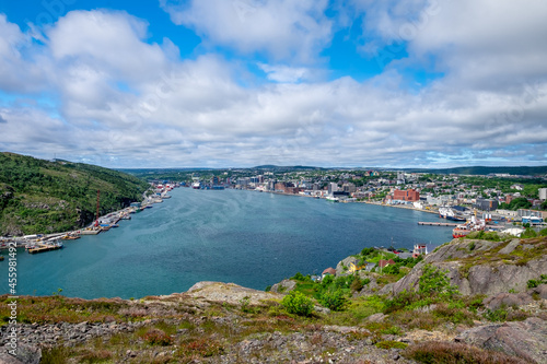 The hillside of St. John's Harbour, Newfoundland, on a sunny day, under blue sky and white clouds. The colorful wooden houses are scattered along the hillside with the blue ocean in the foreground.