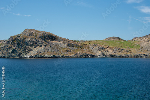 A large rolling hill at the edge of the ocean. The mountain is covered in trees and has a steep rock cliff. The deep blue water is smooth. There's a hiking trail along the edge of the rocky seashore.