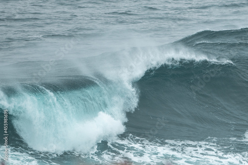 An angry turquoise green colour massive rip curl of a wave as it barrels rolls along the ocean. The white mist and froth from the wave are foamy and fluffy. The ocean in the background is deep blue. 