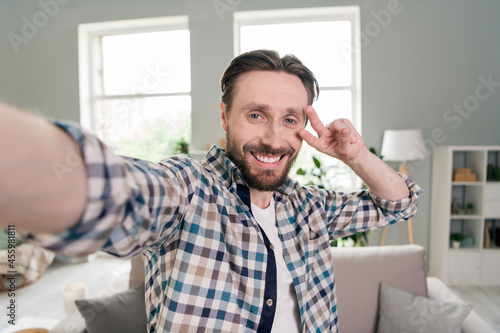 Self-portrait of attractive cheerful guy showing v-sign near eye good mood staying home at light room indoors