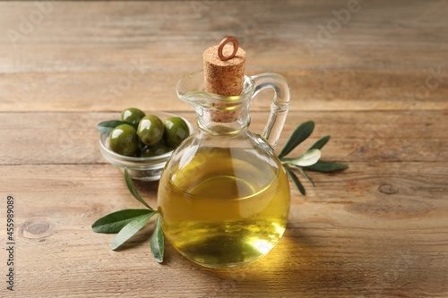 Glass jug of oil, ripe olives and green leaves on wooden table, closeup
