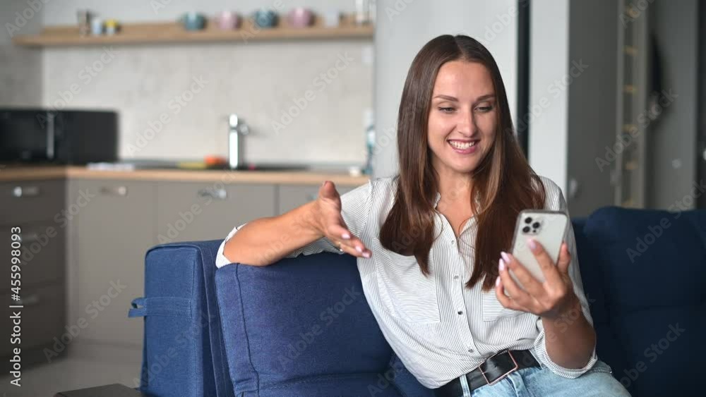 Video connection on the distance. Positive young woman using the smartphone for video call. Smiling female has pleasant online meeting sitting on the couch at home, enjoys online chatting with friends Video connection on the distance. Positive young woman using the smartphone for video call. Smiling female has pleasant online meeting sitting on the couch at home, enjoys online chatting with friends