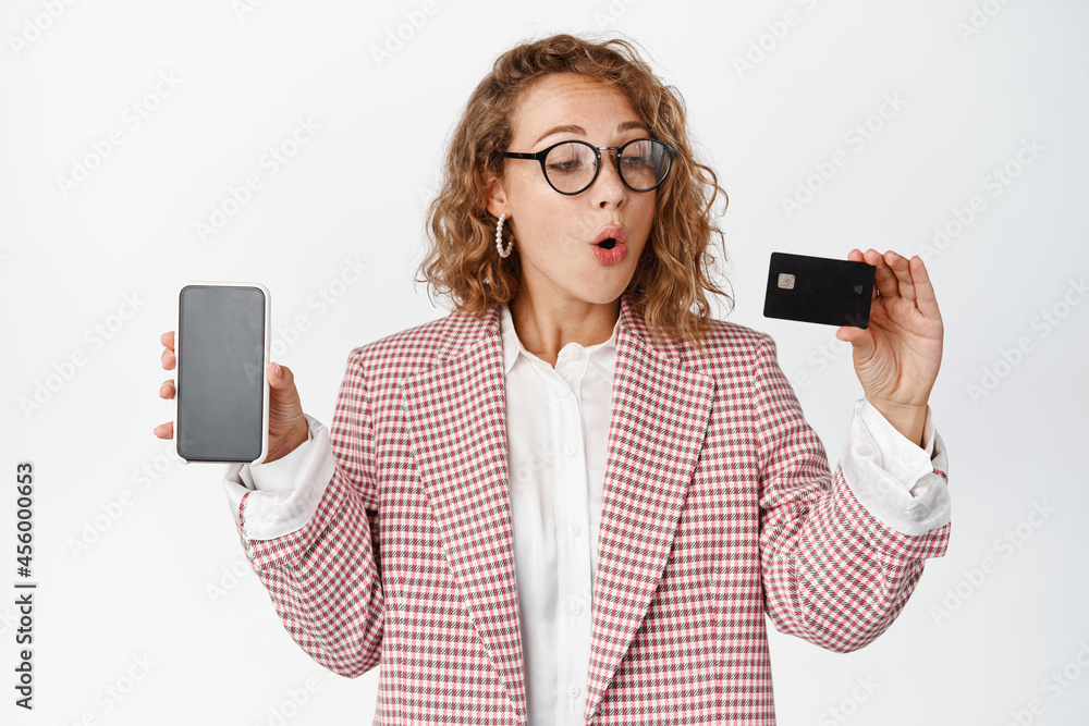 Excited business woman in glasses, shows mobile phone screen, looks at credit card with amazed face expression, white background Excited business woman in glasses, shows mobile phone screen, looks at credit card with amazed face expression, white background