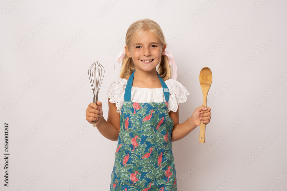 Young smiling girl wearing kitchen apron and holding kitchen utensils over white background Young smiling girl wearing kitchen apron and holding kitchen utensils over white background
