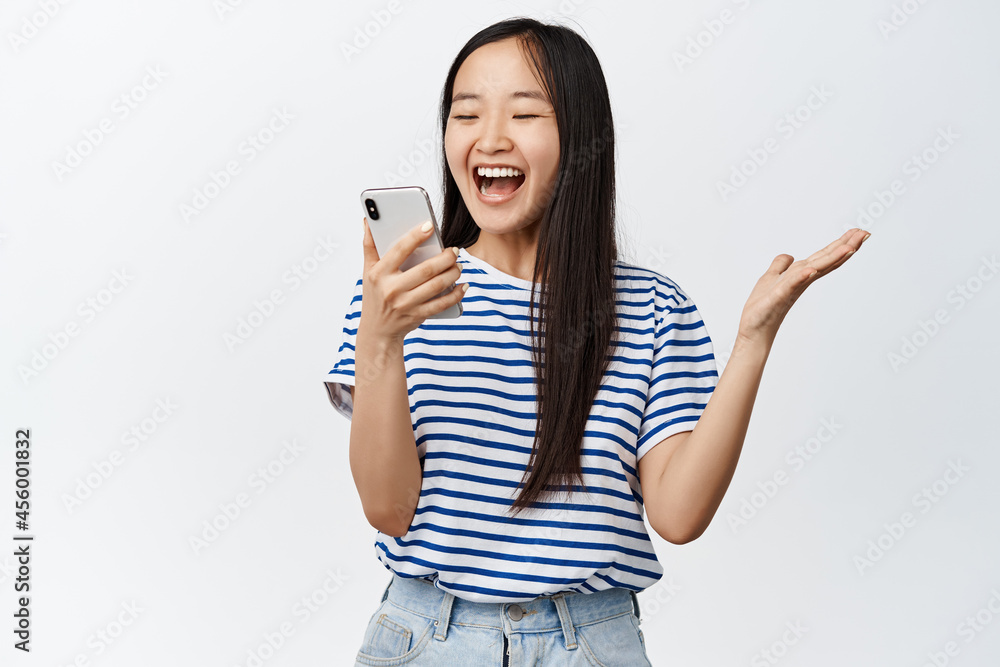 Image of asian brunette girl looking with excited and surprised face at smartphone, reacting amazed at mobile phone notification, standing over white background Image of asian brunette girl looking with excited and surprised face at smartphone, reacting amazed at mobile phone notification, standing over white background