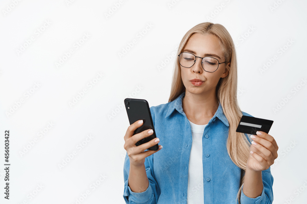 Image of girl in glasses looking thoughtful at smartphone screen, holding credit card, buying something, making decision while online shopping, standing over white background Image of girl in glasses looking thoughtful at smartphone screen, holding credit card, buying something, making decision while online shopping, standing over white background
