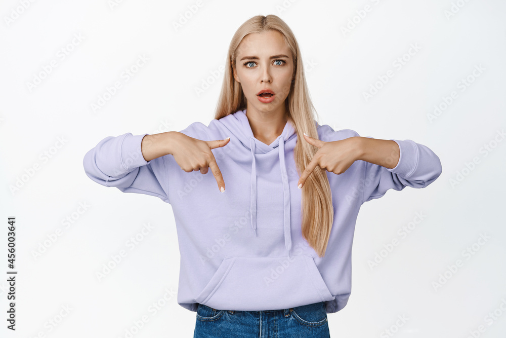 Shocked blond girl pointing fingers down, looking startled at camera, standing in casual clothes over white background Shocked blond girl pointing fingers down, looking startled at camera, standing in casual clothes over white background