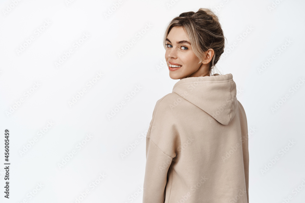 Young stylish woman looking behind her shoulder and smiling happy, standing in beige hoodie over white background Young stylish woman looking behind her shoulder and smiling happy, standing in beige hoodie over white background