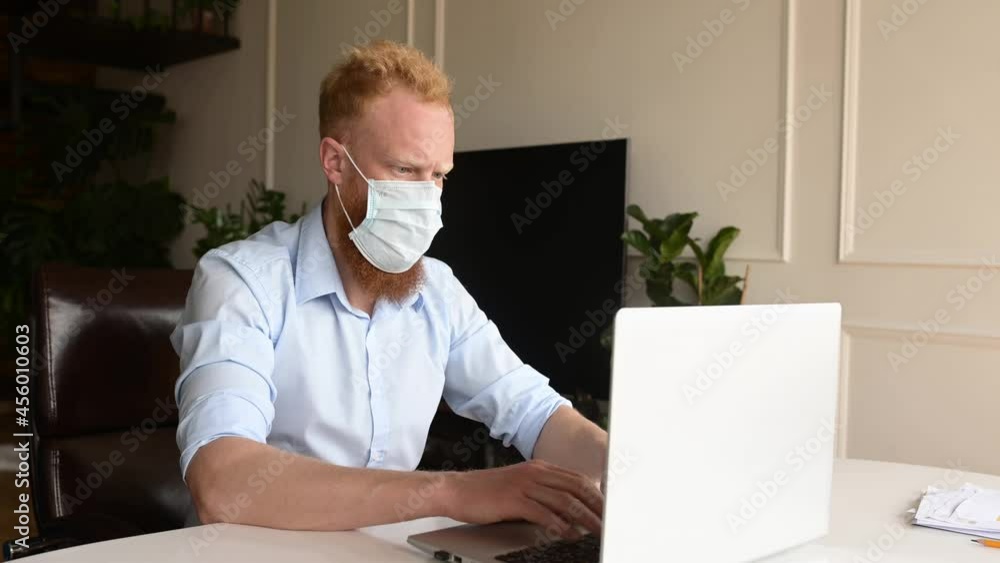 Worried redhead bearded man wearing protective medical mask working with a lalptop in office, pensive man sits at the desk resting chin on hand, feels melancholy Worried redhead bearded man wearing protective medical mask working with a lalptop in office, pensive man sits at the desk resting chin on hand, feels melancholy