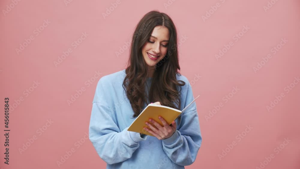 A smiling brunette woman coming up with an idea while writing it on her notebook standing isolated over pink background in the studio A smiling brunette woman coming up with an idea while writing it on her notebook standing isolated over pink background in the studio
